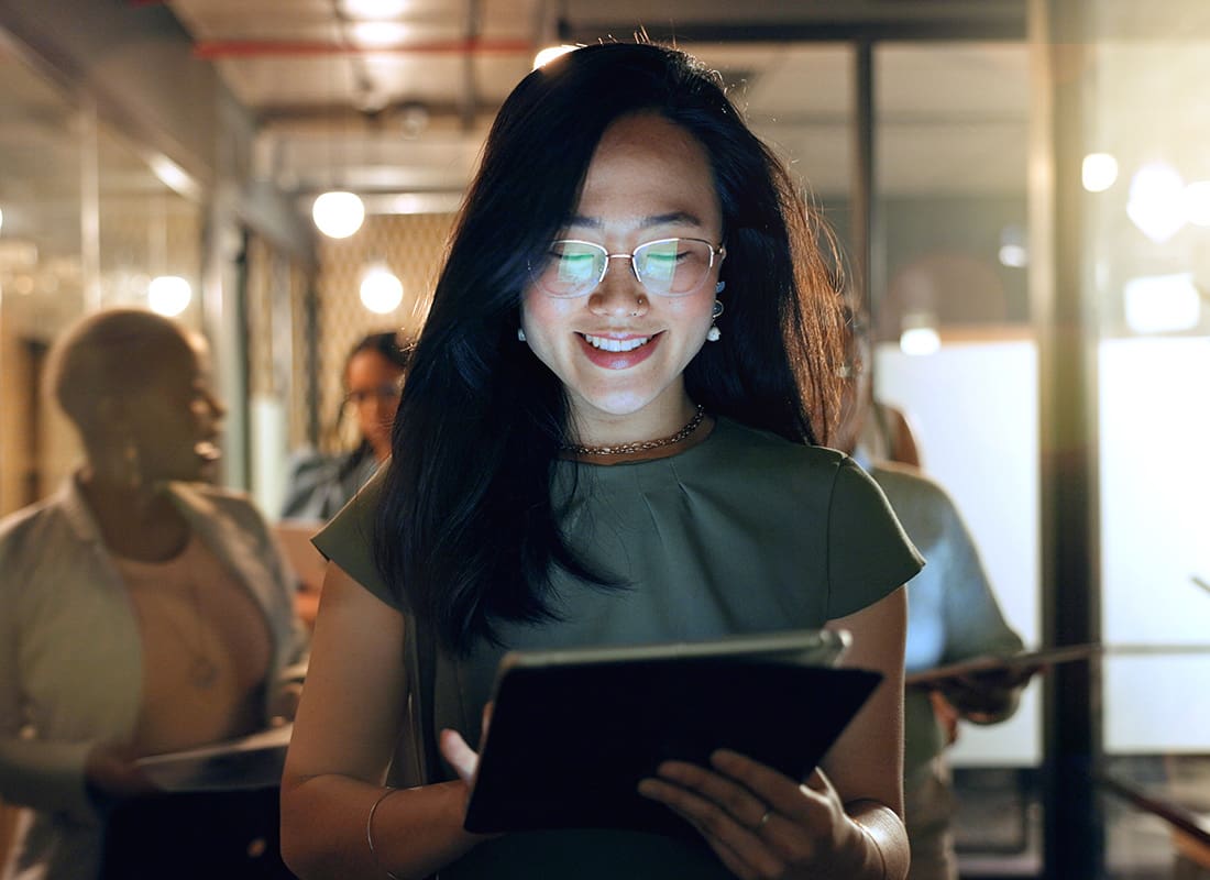 Service Center - Closeup Portrait of a Smiling Young Asian Business Woman Looking at a Tablet While Walking Through a Hallway in the Office with Other Employees Behind Her